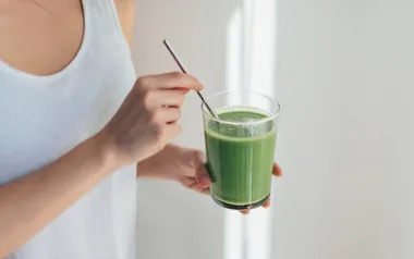 Person in white tank top holding a glass of green smoothie with a metal straw.