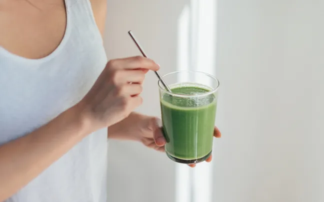 Person in white tank top holding a glass of green smoothie with a metal straw.