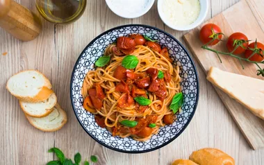 Bowl of spaghetti with tomato sauce and basil, surrounded by bread, cheese, tomatoes, and olive oil on a wooden table.