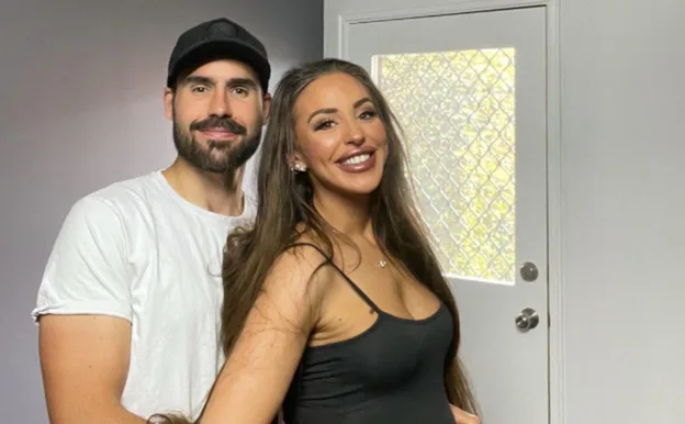 A smiling couple stands together indoors, with the woman in a black top and the man in a white shirt and cap.