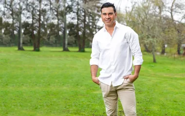 Man in white shirt and beige pants standing outdoors with hands in pockets, in front of a green grassy field and trees.