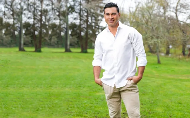 Man in white shirt and beige pants standing outdoors with hands in pockets, in front of a green grassy field and trees.