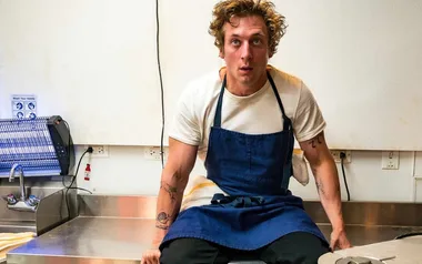 A person wearing a blue apron sitting in a kitchen, looking surprised.