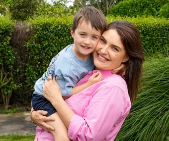 Woman in pink shirt holding and smiling with a young boy, outdoors with greenery in the background.