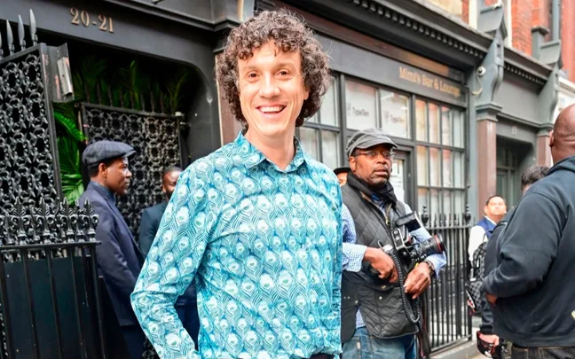A person in a patterned shirt smiles outside a building with "Mimi's Bar & Lounge" sign, surrounded by a group.