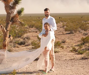 Couple dressed in white poses beside a tree in a desert landscape, with a flowing sheer fabric.