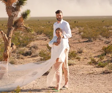 Couple dressed in white poses beside a tree in a desert landscape, with a flowing sheer fabric.