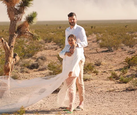Couple dressed in white poses beside a tree in a desert landscape, with a flowing sheer fabric.