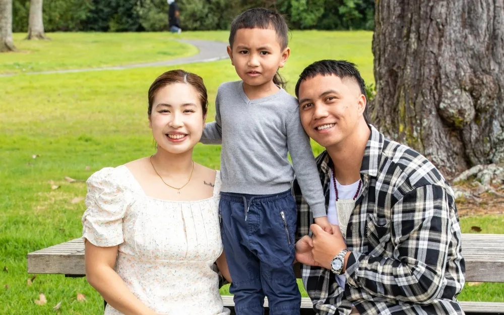 Family sitting on a park bench, smiling. Woman in white dress, child in gray top, man in checkered shirt.