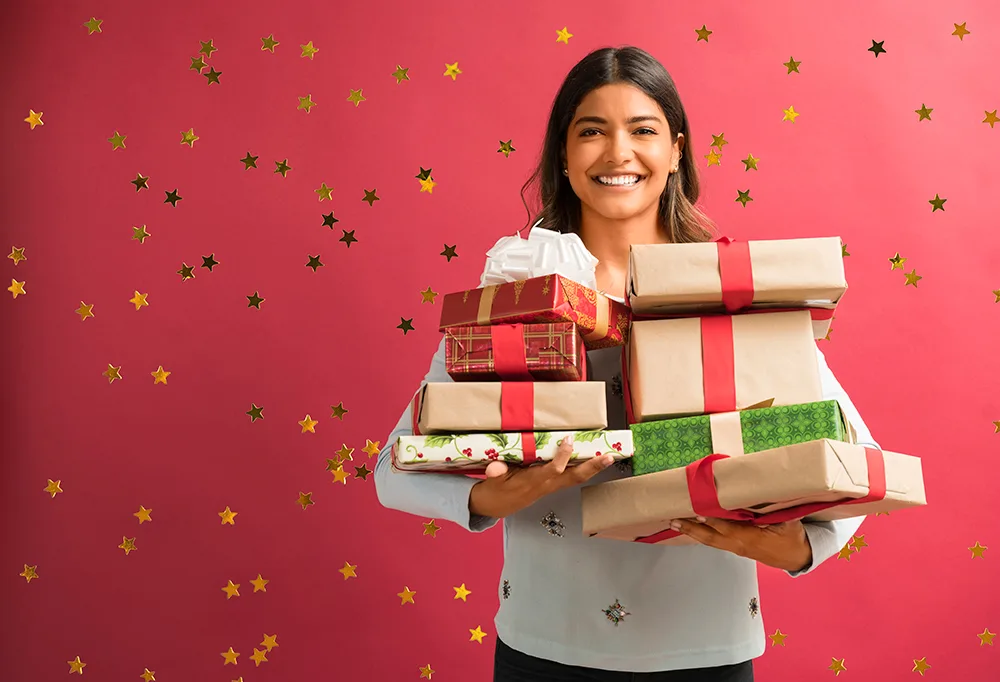 Smiling person holding stacked gift boxes with red ribbons on a starry red background.