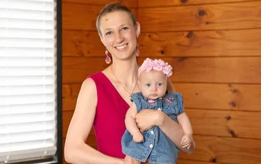 Woman in a pink dress holding a baby wearing a denim outfit and pink bow, in a wooden room.