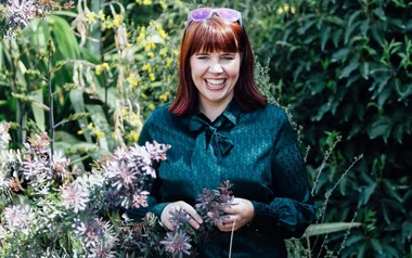 A woman with red hair and sunglasses stands smiling in a garden, surrounded by green foliage and purple flowers.