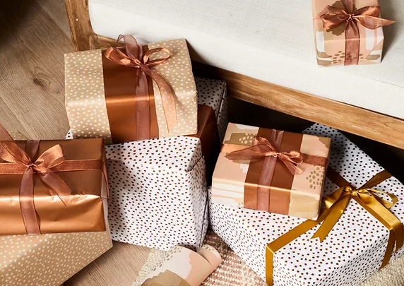 Gift boxes wrapped in brown and white patterned paper with bows, placed on a wooden floor by a white bench.