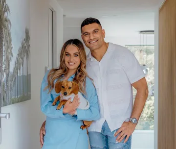 A smiling couple poses inside a bright hallway, with the woman holding a small dog.