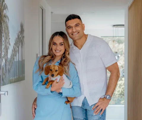 A smiling couple poses inside a bright hallway, with the woman holding a small dog.