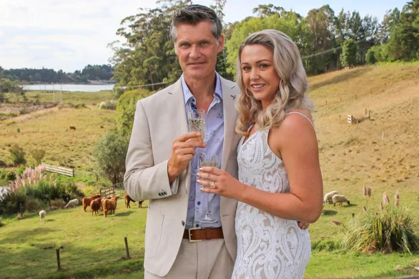A couple in formal attire holds drinks outside, with a rural landscape and livestock in the background.