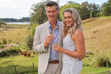 A couple in formal attire holds drinks outside, with a rural landscape and livestock in the background.