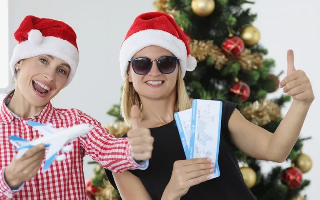 Two women in Santa hats show thumbs up with plane tickets and a model airplane near a decorated Christmas tree.
