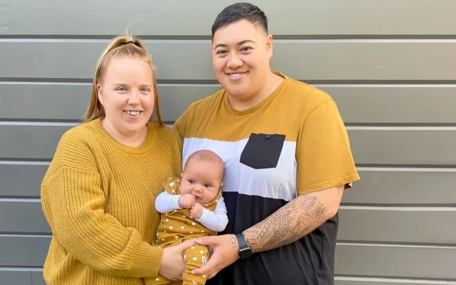 A couple smiles while holding a baby, all wearing mustard-colored clothing, against a striped wall background.