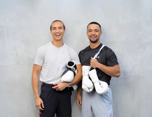 Two smiling men with boxing gloves stand against a gray wall.