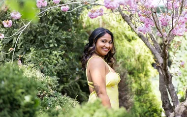 Smiling woman in a yellow dress stands among green bushes and pink flowering trees.