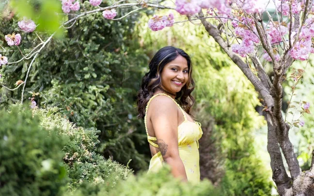 Smiling woman in a yellow dress stands among green bushes and pink flowering trees.