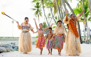 Pacific Island family in traditional attire joyfully dancing on a tropical beach.