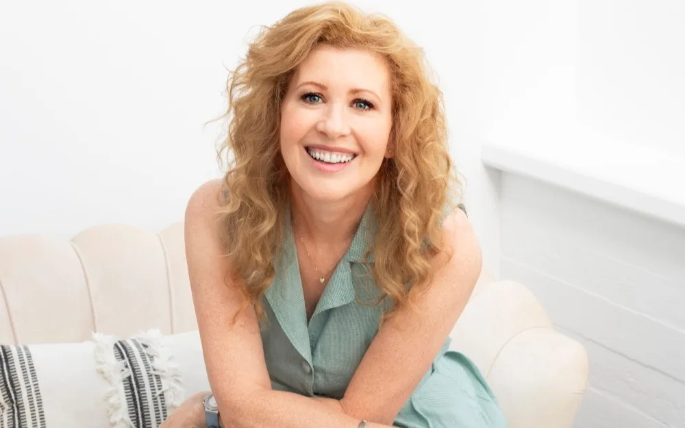 A woman with curly blonde hair smiling, seated on a light-colored sofa in a bright room.