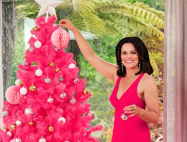Woman in pink dress decorates a bright pink Christmas tree with ornaments in a room with large windows.
