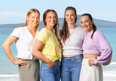 Four women standing together on a beach, smiling at the camera with the ocean and distant hills in the background.