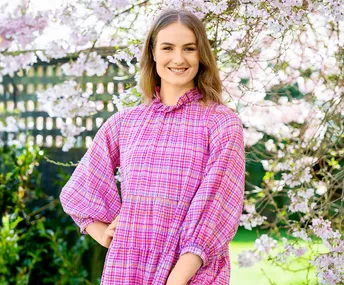 A person in a pink checked dress stands smiling in front of blooming cherry blossoms.