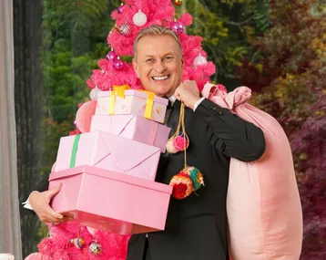 Man in suit holding pink gift boxes and a sack, standing by a pink Christmas tree.