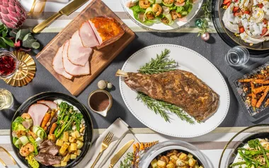 Festive table setting with sliced ham, roast lamb, salads, sweet potatoes, and desserts, garnished with holiday decor.