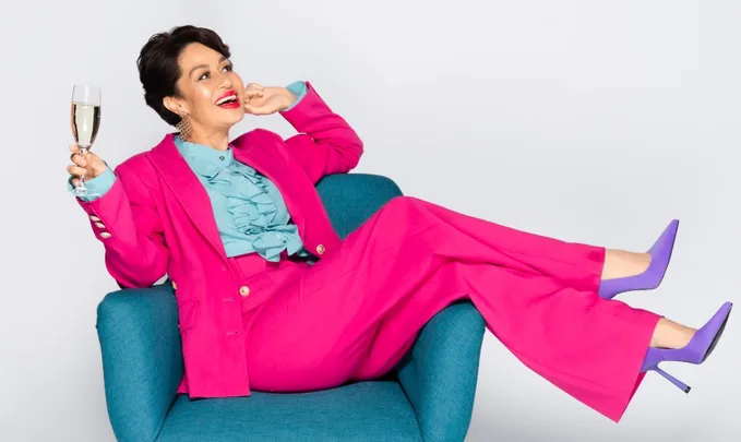 Woman in a vibrant pink suit relaxing on a chair, holding a champagne glass, and wearing purple heels.