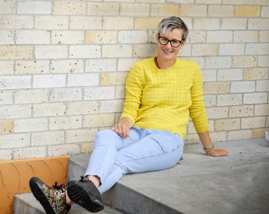 Person in a yellow sweater and jeans sitting against a brick wall, smiling.