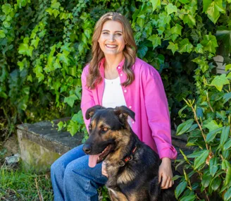 A woman in a pink shirt smiles while sitting with a black and brown dog in front of lush green foliage.
