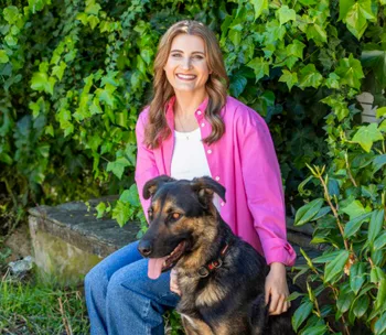 A woman in a pink shirt smiles while sitting with a black and brown dog in front of lush green foliage.