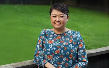A person smiling in a floral blouse stands in front of a green grassy background.