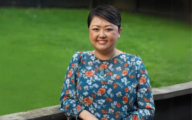 A person smiling in a floral blouse stands in front of a green grassy background.