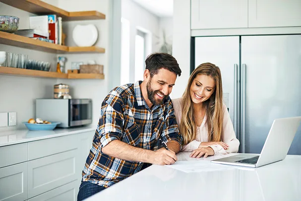 A man and woman smiling while working on paperwork together at a kitchen counter, with a laptop nearby.