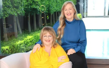Two smiling women sit together, one in a yellow top and the other in blue, with a green garden and pool in the background.