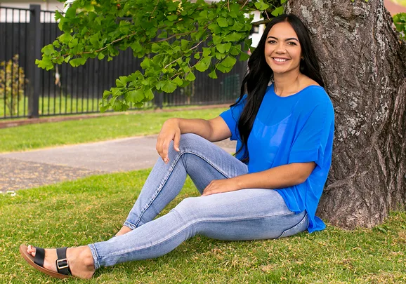A woman in a blue top and jeans sits smiling under a tree in a park.