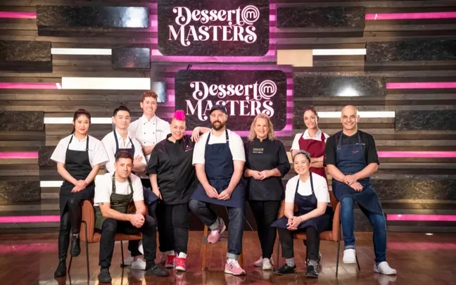 Group of chefs posing in front of "Dessert Masters" sign, wearing aprons, smiling for the camera.