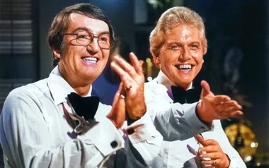 Two entertainers in white shirts and bow ties smile and gesture enthusiastically.