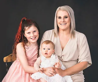 A smiling woman with blonde hair holds a baby, next to a young girl with red hair, all wearing light dresses, against a dark backdrop.