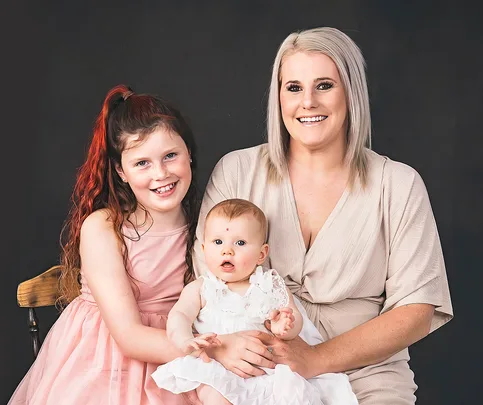 A smiling woman with blonde hair holds a baby, next to a young girl with red hair, all wearing light dresses, against a dark backdrop.