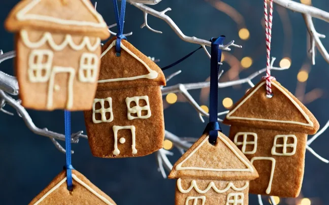 Gingerbread cookies shaped like houses, decorated with white icing, hanging on a white branch with colorful ribbons.