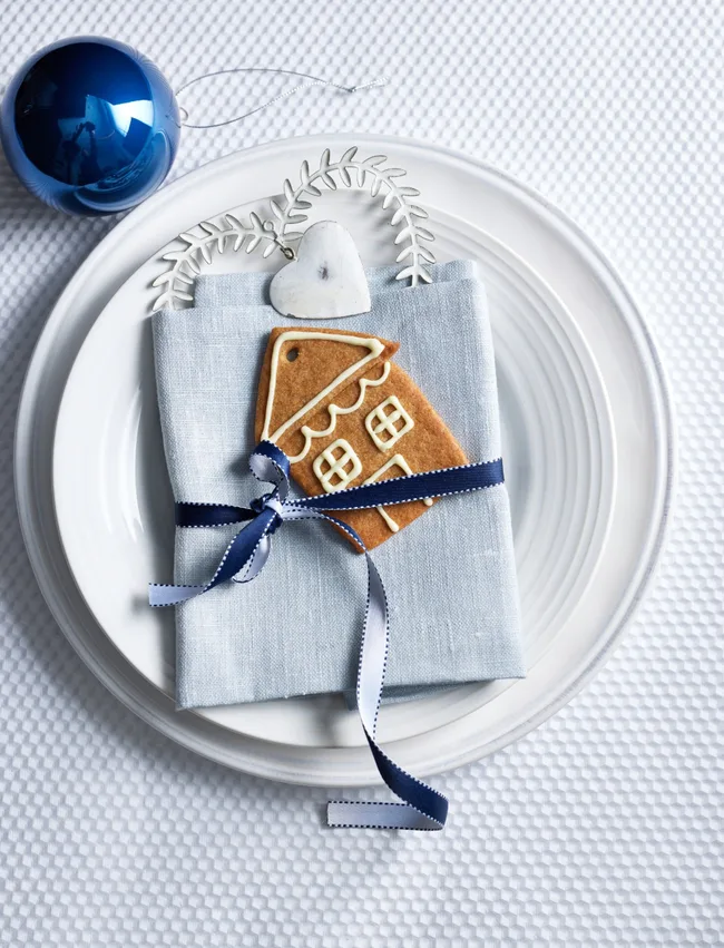 Plate with blue napkin, gingerbread house cookie, tied ribbon, and heart ornament, beside a blue bauble on white cloth.