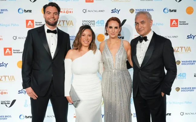 A group of four people in formal attire posing on the red carpet at an event with a logo backdrop.