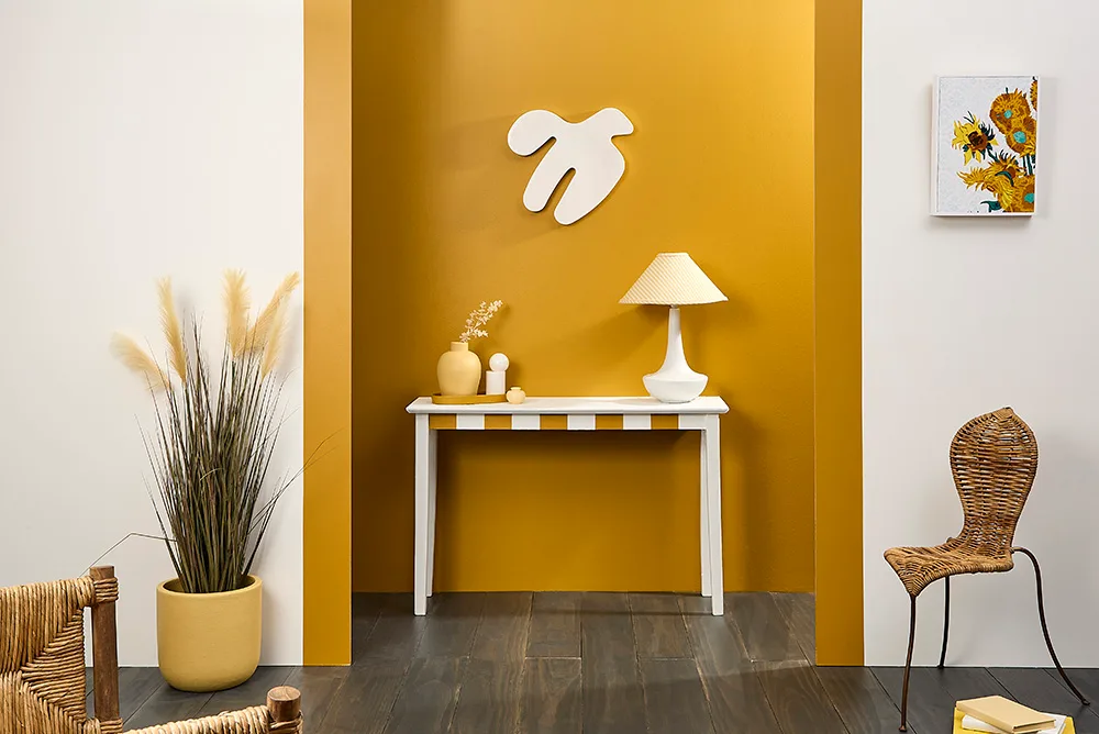 Stylish interior with a white console table, lamp, wall art, and wicker chair against a mustard yellow wall.